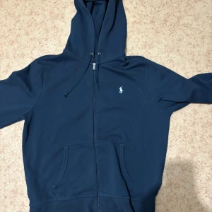 Marinblå hoodie från Polo Ralph Lauren - Snygg marinblå hoodie från Polo Ralph Lauren med dragkedja och huva. Klassisk logga broderad på bröstet, praktisk känguruficka och snörning i huvan. Perfekt för chill dagar eller när du vill ha en clean streetstyle-look.