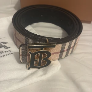 Burberry rutigt bälte med svartspänne - Snyggt bälte från Burberry med klassiskt rutig design i beige, svart och rött. Bältet har ett stort, guldigt spänne med Burberrys ikoniska B-logga. Tillverkat i skinn och levereras med originalpåse och box. Perfekt för att lyfta vilken outfit som helst.