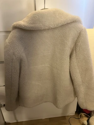 Beige teddyjacka från NA-KD, strl 36 - Supermysig teddyjacka från NA-KD i beige färg med stora svarta knappar och bred krage. Jackan har två rejäla fickor framtill och är helfodrad för extra värme. Perfekt för dig som vill ha en chill och trendig look under höst och vinter.