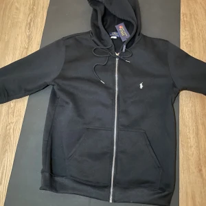 Svart hoodie från Polo Ralph Lauren - Svart hoodie från Polo Ralph Lauren med dragkedja och snörning i huvan. Klassisk design med vit broderad logga på bröstet och stora fickor framtill. Tillverkad i mjuk bomull, perfekt för chill dagar.