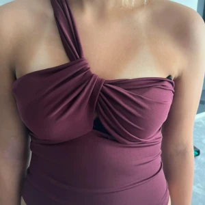 Vinröd oneshoulder topp med twist - Säljer en vinröd topp med snygg twistad detalj framtill och oneshoulder-design. Toppen är figurnära och har ett coolt draperat tyg över bysten. Materialet känns mjukt och stretchigt, perfekt för dig som vill sticka ut med en unik look.