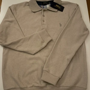 Beige pikétröja från Polo Ralph Lauren - Snygg beige pikétröja från Polo Ralph Lauren med klassisk krage och knappar framtill. Tröjan har långärmade ribbade muddar och den ikoniska broderade loggan på bröstet. Perfekt för dig som gillar stilrena och tidlösa plagg.