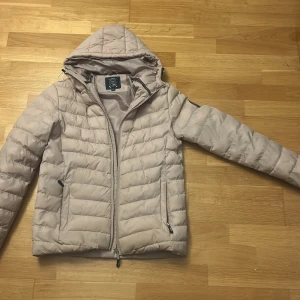 Beige pufferjacka från Vinson - Snygg beige pufferjacka från Vinson med luva och två sidofickor med dragkedja. Jackan har quiltad design och ett rundat märke på ärmen. Perfekt för kyliga dagar och har en clean look som funkar till många stilar.