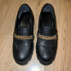 Svarta chunky loafers med guldkedja - Snygga svarta loafers med chunky platt sula och dekorativ guldfärgad kedja över foten. Skorna är från justfab och är i storlek 38. Dom är använda några gånger och är lite nötta på sulan men det är inget man tänker på om man inte kollar nära