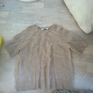 Beige stickad tröja från NA-KD - Mysig beige stickad tröja från NA-KD i storlek S. Tröjan har korta ärmar och breda ribbade muddar vid hals, ärmslut och nederkant. Perfekt för lager-på-lager och chill stil. Enkel design som funkar till många looks.