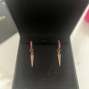 Guldörhängen från L8Hasselblad - Snygga hängande örhängen i gulddoublé med nitar med infattade rosa zirkoner både på ringen och taggen. Ger en cool och trendig vibe, perfekt för dig som gillar att sticka ut med färg och form. Passar till flera piercings och kan kombineras med andra smycken. Säljer då jag bär silver numera. Nypris: 1000 kr par, l8hasselblad ask ingår 