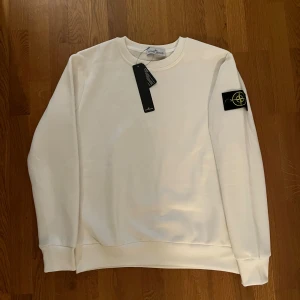 Vit sweatshirt från Stone Island - Vit sweatshirt från Stone Island med klassisk rund hals och ribbade muddar. Tröjan har lång ärm och den ikoniska Stone Island-loggan på vänster ärm. Tillverkad i mjuk bomull för skön komfort. Perfekt för dig som gillar stilrena och cleana plagg. Lite stor i storlek.