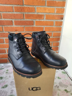 Svarta UGG boots i skinn - Säljer ett par svarta UGG boots med snörning och robust platt sula. Skorna är tillverkade i skinn med detaljer i mörk mocka runt skaftet. UGG-logga bak på hälen och vattentäta egenskaper. Perfekta för höst och vinter, riktigt snygga och stilrena.