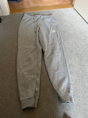 Grå mjukisbyxor från Nike, storlek S - Grå mjukisbyxor från Nike med klassisk logga på vänster lår. Byxorna har snörning i midjan och muddar vid bensluten för en skön passform. Perfekta för chill eller träning, tillverkade i mjukt bomullsmaterial.