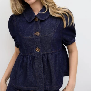 Mörkblå denimskjorta med puffärm - Supersöt mörkblå skjorta i denim med puffiga korta ärmar och stora bruna knappar framtill. Skjortan har en rund krage och en volangliknande söm vid midjan som ger en cool och avslappnad siluett. Perfekt att styla med shorts eller jeans för en chill look.
