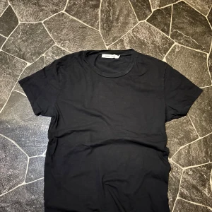 Svart t-shirt från Samsøe Samsøe - En stilren svart t-shirt från Samsøe Samsøe i mjuk bomull. Klassisk rund hals och korta ärmar, perfekt för en clean och enkel look. Passar dig som gillar minimalistisk stil och vill ha ett plagg som funkar till allt.