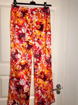 Blommiga vida byxor från Amisu - Färgglada vida byxor från Amisu i storlek 42 med stort blommigt mönster i orange, rött, rosa, gult och vitt. Byxorna har elastisk midja och är tillverkade i ett lätt, glansigt material som känns skönt mot huden. Perfekt för dig som gillar att sticka ut.