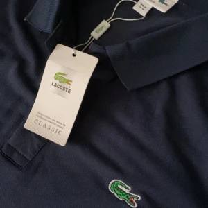 Marinblå pikétröja från Lacoste - Snygg marinblå pikétröja från Lacoste med klassisk krage och knappar framtill. Ikoniska gröna krokodilloggan är broderad på bröstet. Pikén är i toppskick…bara att höra av sig vid funderingar🤝🫡