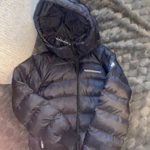 Svart BARNVINTERJACKA strlk 130 Peak Performance - Säljer en svart barnvinterjacka från Peak Performance med huva och dragkedja framtill. Jackan har ett quiltat mönster och är långärmad. Perfekt för kalla dagar och har en clean, sportig look med diskret logga på bröstet. Storlek 130