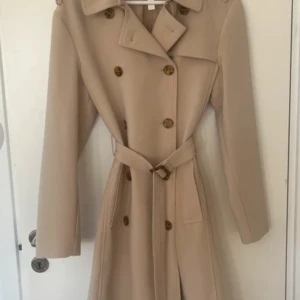 Beige trenchcoat från H&M med bälte - Snygg beige trenchcoat från H&M med klassisk dubbelknäppning och bälte i midjan. Jackan har långa ärmar, bred krage och stilrena knappar i sköldpaddsmönster. Perfekt för dig som gillar clean och tidlös stil.