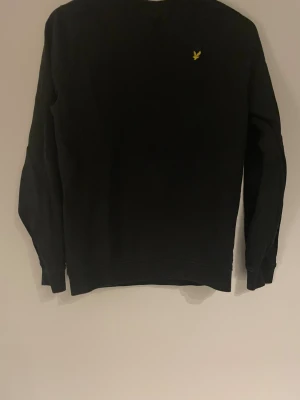 Svart sweatshirt från Lyle & Scott - Svart sweatshirt från Lyle & Scott med klassisk rund hals och långärmad passform. Tröjan har en gul broderad logga på bröstet och är tillverkad i mjuk bomull. Perfekt för dig som gillar stilrena och enkla plagg med en touch av street.