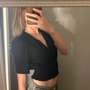 Svart croppad blus med v-ringning - Säljer en svart croppad blus med v-ringning och stora klädda knappar framtill. Toppen har korta ärmar och smockad midja som ger en snygg passform. Perfekt till jeans eller kjol för en trendig look. 