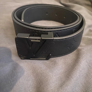 Svart L V skärp i skinn - Snyggt svart skärp från L V med klassisk L V-logga som spänne. Bältet är gjort i skinn och har präglade monogramdetaljer längs hela bandet. Perfekt accessoar för att lyfta din outfit med en lyxig touch.