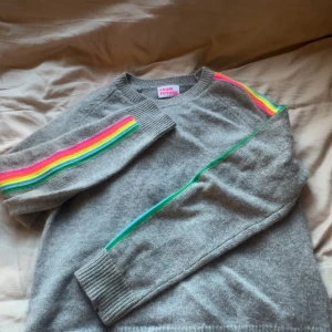 Grå kashmir tröja med regnbågsränder - Grå stickad tröja från From Future med färgglada regnbågsränder längs ärmarna i rosa, orange, gul, blå och grön. Tröjan är i helt nytt skick.
