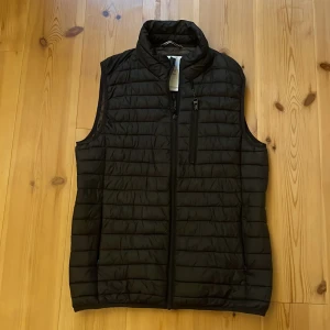 Svart dunväst från Esprit, storlek M - Svart quiltad dunväst från Esprit med hög krage och dragkedja framtill. Västen har två sidofickor med dragkedja och en bröstficka med dragkedja. Stilren design med horisontella sömmar och lätt vaddering, perfekt för lager-på-lager under kyliga dagar. Använd relativt mycket men ändå i bra skick