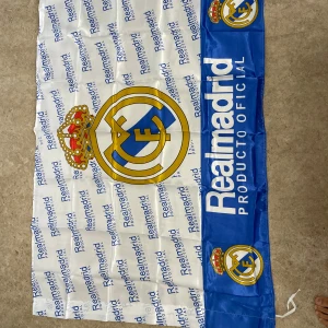 Real Madrid halsduk i blått och vitt - Stor halsduk med Real Madrid-logga och texten 'Producto Oficial' tryckt i blått och vitt. Halsduken har klubbens emblem i gult, rött och blått samt upprepade loggor och text över hela ytan. Perfekt accessoar för alla fotbollsfans.