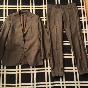 Brun kostym från Rose & Born, stl 48 - Snygg brun kostym från Rose & Born i italiensk design. Kavajen har två knappar, klassiska fickor och smal passform. Byxorna har pressveck och uppvikta benslut. Tillverkad i 100% ull för en lyxig känsla. Passar perfekt för dig som är mellan 180-183 i storlek. Perfekt för dig som vill ha en stilren och tidlös look. 
