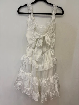 Vit volangklänning med rosett - Supersöt vit kortklänning med flera lager volanger och smock upptill. Axelbanden är dekorerade med volanger och framtill finns en stor rosett som detalj. Klänningen har en romantisk och lekfull vibe, perfekt för sommarens alla tillfällen.