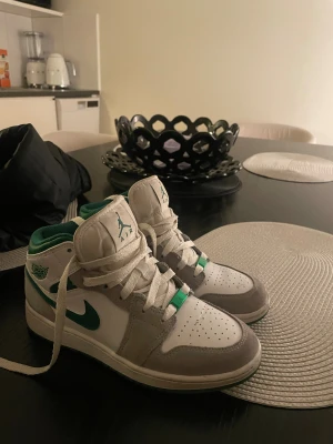 Nike Air Jordan 1 Mid vit/grön - Nike Air Jordan 1 Mid sneakers i vitt, grått och grönt med klassisk snörning och rund tå. Ovandelen är i skinn och mocka, med gröna detaljer på swoosh och logga. Skön platt sula och ikonisk Jordan-vibe. Perfekt för dig som gillar streetstyle.