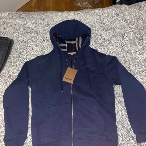 Marinblå Burberry hoodie  - Snygg marinblå hoodie från Burberry med klassiskt rutigt foder i huvan och dragkedja framtill. Tröjan har snörning vid huvan, två fickor och ribbade muddar. Perfekt för dig som gillar stilrena märkesplagg med coola detaljer.