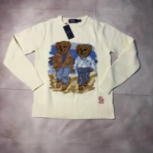 Vit stickad tröja Polo Ralph Lauren - Vit stickad tröja från Polo Ralph Lauren med två broderade björnar på framsidan, RL 85 broderat i rött vid nederkanten. Tröjan har rund hals och långa ärmar, ribbade muddar och är tillverkad i mjuk bomull. Perfekt för dig som gillar ikoniska motiv och klassisk stil.