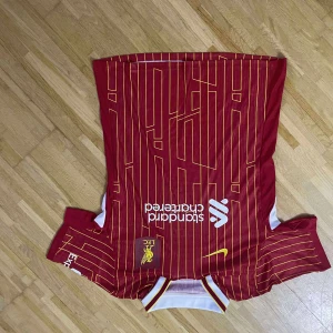 Liverpool FC röd randig fotbollströja - Säljer en officiell Liverpool FC fotbollströja från Nike i rött med gula ränder och vita detaljer. Tröjan har korta ärmar, klubbmärke och sponsortryck på bröstet. Materialet är lätt och andas, perfekt för match eller träning. Snygg design med modern känsla.