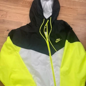 Nike vindjacka med huva, M - Nike vindjacka i svart, vit och neon-gul med huva och hel dragkedja framtill. Jackan har meshfoder, snörning vid halsen och klassisk Nike-logga på bröstet. Perfekt för dig som gillar sportig stil och vill sticka ut med färg.