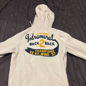 Vit hoodie från Abercrombie & Fitch - Vit hoodie med dragkedja från Abercrombie & Fitch. Broderade detaljer i blått och gult framtill och på ryggen med text och siffror. Huva med snörning och två fickor framtill. Perfekt för en avslappnad och sportig stil.