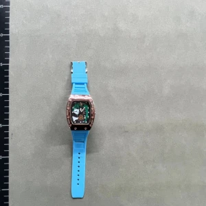 Richard Mille blå klocka med roséguld - Säljer en Richard Mille klocka med cool blå silikonrem och en rektangulär urtavla med roséguldfärgad ram. Urtavlan har synliga mekaniska detaljer och en unik design. Perfekt för dig som gillar att sticka ut med accessoarer. Box medföljer.