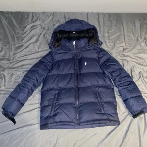 Mörkblå dunjacka från Polo Ralph Lauren - Snygg mörkblå dunjacka från Polo Ralph Lauren med huva och dragkedja framtill. Jackan har det klassiska Polo-logot på bröstet, två sidofickor med dragkedja och justerbara muddar. Perfekt för kalla dagar. Jackan är använd och är storlek L för barn och passar folk mellan 160-170 jackan är i använt skick