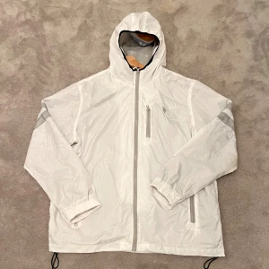 Ralph Lauren Windbreaker Jacka - Vit Ralph Lauren jacka med reflexer på ärmarna. Riktigt bra skick, använd en gång. Enda defekten är att resårbandet har gått av på ena sidan (skriv för bild) men påverkar inte passformen.