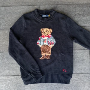 Mörkblå stickad tröja Polo Bear - Mörkblå stickad tröja från Polo Ralph Lauren med ikonisk Polo Bear broderad framtill. Tröjan har rund hals, ribbade muddar och lång ärm. Perfekt statement-piece för dig som gillar klassisk stil med en twist.