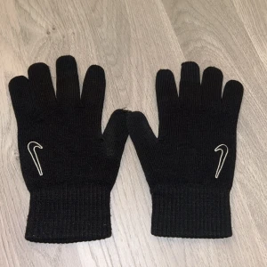 Svarta stickade Nike handskar - Svarta stickade handskar från Nike med klassisk vit logga på ovansidan av varje handske. Perfekt för kalla dagar och snyggt till streetwear. Ribbstickade muddar för extra komfort och passform.
