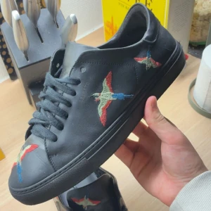 Svarta sneakers från Axel Arigato - Svarta sneakers i skinn från Axel Arigato med broderade fåglar i rött, blått och beige på sidorna och tån. Skorna har svarta snören och en klassisk låg profil. Perfekta för dig som vill ha en stilren men unik look.