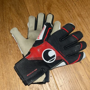 Svarta och röda målvaktshandskar Uhlsport - Säljer ett par målvaktshandskar från Uhlsport med svart och röd design, vita detaljer och logga på ovansidan. Lite använda men funkar fortfarande bra. Handflatan är ljusgrå och har ett slitstarkt material för bra grepp. Handskarna har justerbar kardborre vid handleden och coola röda linjer på ovansidan. Nypris 700kr ish