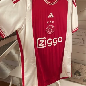 Ajax fotbollströja Adidas röd/vit L - Säljer en snygg och klassisk Ajax-tröja i storlek L . Tröjan är i mycket bra skick utan fläckar eller skador. Perfekt för dig som samlar på matchtröjor, tränar i kvalitetsplagg eller bara gillar Ajax ikoniska stil. Priset är sänkbart vid snabb affär