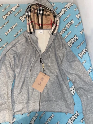 Grå Burberry hoodie med rutigt foder - Snygg grå hoodie från Burberry i 100% bomull. Klassisk dragkedja framtill, snörning i huvan och ikoniskt rutigt foder i huvan. Broderad Burberry-logga på bröstet och praktisk känguruficka. Perfekt för en stilren och avslappnad look.
