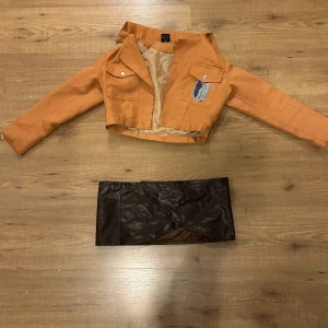 Attack on Titan cosplay - Säljer en croppad orange jacka inspirerad av Attack on Titan med broderade emblem på bröstet, ryggen och ärmen. Även underdel. Stl S