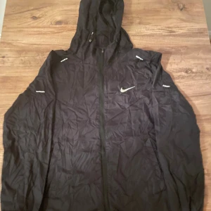 Svart vindjacka från Nike med huva - Svart vindjacka från Nike med huva och hel dragkedja framtill. Jackan har flera vita Nike-loggor på bröstet och ärmarna, samt diskreta reflexdetaljer. Materialet är lätt och syntetiskt, perfekt för blåsiga dagar. Jackan har en sportig och clean look.