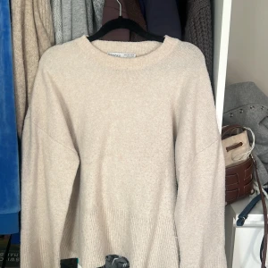 Beige stickad tröja från Bershka - Mysig beige stickad tröja från Bershka i storlek S. Tröjan har rund hals, ribbade muddar och långärmad passform. Perfekt för lager-på-lager och chill dagar. Enkel och stilren design som funkar till jeans eller kjol.