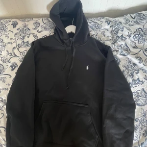 Svart hoodie från Polo Ralph Lauren - Svart hoodie från Polo Ralph Lauren med klassisk logga broderad på bröstet. Tröjan har huva med snörning och en stor magficka framtill. Mjuk bomullskvalitet och långärmad modell som är perfekt för chill dagar.