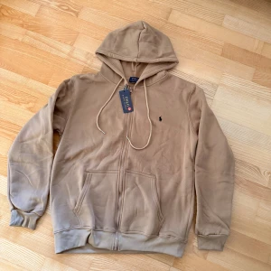 Beige hoodie från Polo Ralph Lauren - Snygg beige hoodie från Polo Ralph Lauren i storlek M. Tröjan har huva med snörning, dragkedja framtill och två stora fickor. Klassisk broderad logga på bröstet och ribbade muddar vid ärmslut och nederkant. Perfekt för chill dagar.