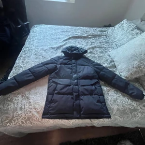 Svart pufferjacka från Jack & Jones - Säljer en svart pufferjacka från Jack & Jones med huva och diskret logga på bröstet. Jackan har långa ärmar och är tillverkad i syntetmaterial med quiltad design. Perfekt för kalla dagar och har en clean, modern look.
