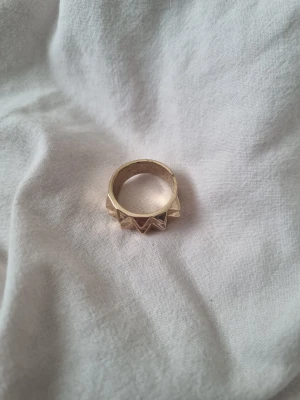 Guldfärgad ring med pyramidmönster - Säljer en cool guldfärgad ring med upphöjda pyramidformade detaljer runt hela ringen. Den har en chunky och edgy vibe som verkligen sticker ut. Perfekt för dig som gillar statement-smycken och vill addera lite attitude till din stil.