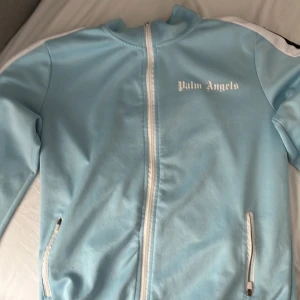 Ljusblå trackjacka från Palm Angels - Säljer en ljusblå trackjacka från Palm Angels med vit dragkedja och vita detaljer längs ärmarna. Jackan har ståkrage, två fickor med dragkedja och logotypen tryckt på bröstet. Perfekt för en chill och sportig stil.